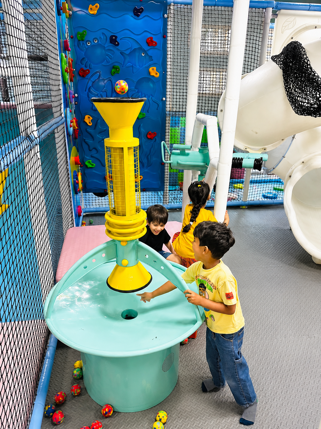 Kids playing at a ball-launcher station with climbing wall and tube slide